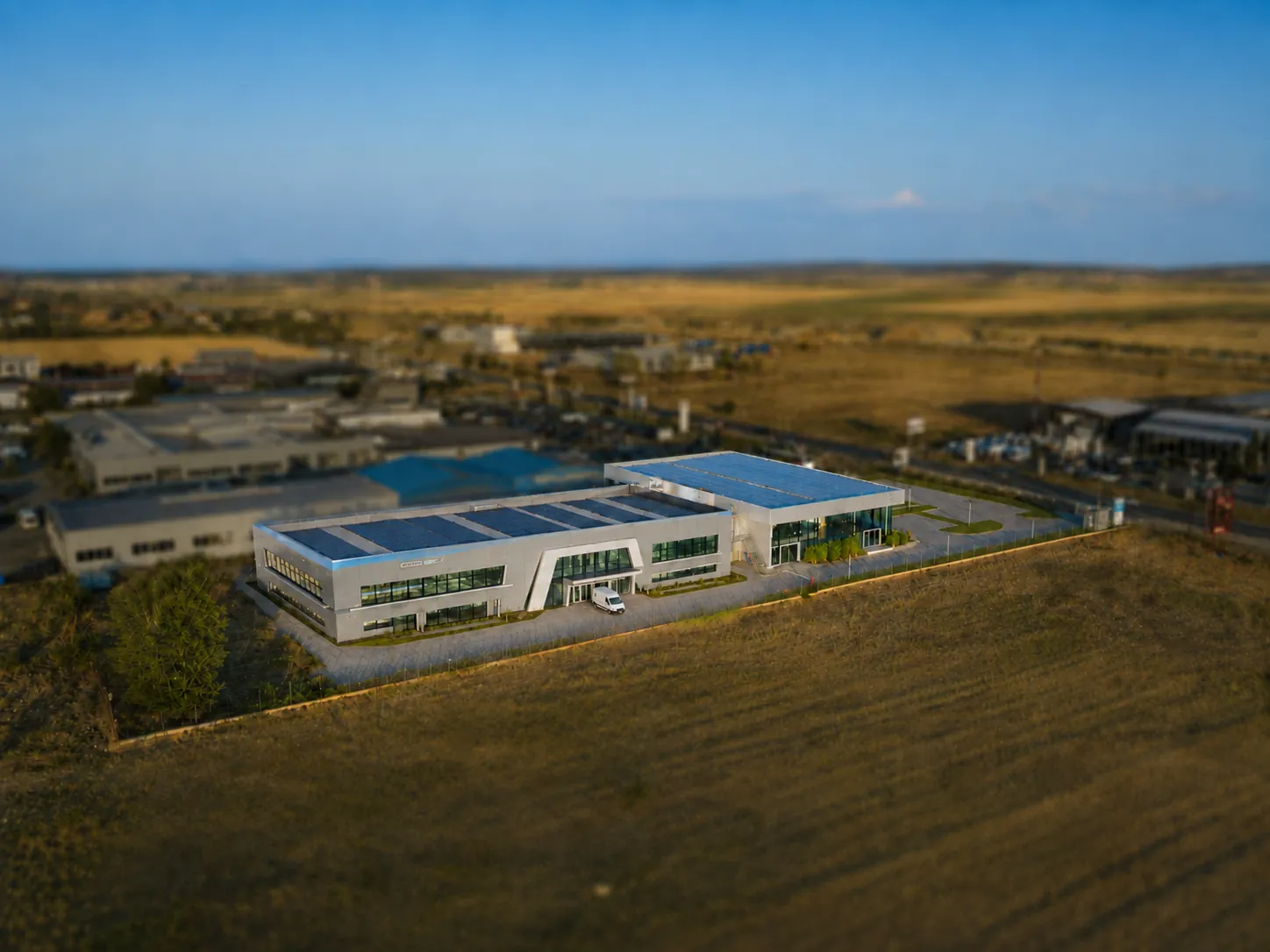 Aerial view of Building B — the hall roofed in solar panels, attached to the glass pavilion — with the surrounding industrial park out of focus