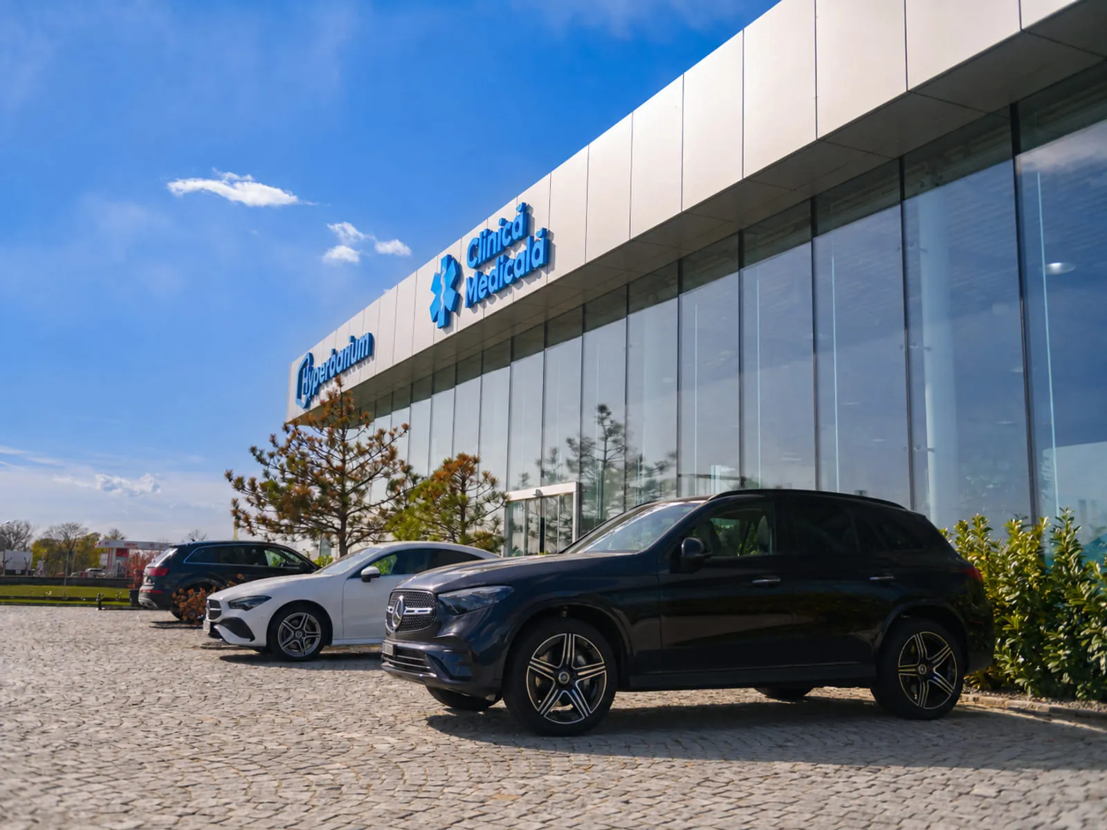 Ground-level view along the glazed clinic frontage, the Hyperbarium and Clinica Medicala signage on the white fascia above cars parked on the paved forecourt